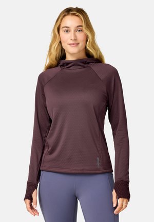 Kvinde iført en langærmet vinrød hættetrøje og blå leggings, stående mod en ensfarvet lys baggrund.