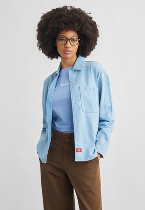 Ung kvinde med stort krøllet hår og briller iført en lyseblå skjorte over en blå t-shirt og brune bukser, hænderne i lommerne.