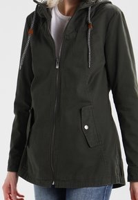 Femme portant une veste à capuche zippée vert foncé avec cordons de serrage et poches à rabat, par-dessus une chemise blanche et un jean bleu.