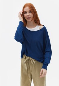Blauwe oversized trui met brede halslijn en geribbelde manchetten, gecombineerd met beige hoge taillebroek die bij de taille is gestrikt.