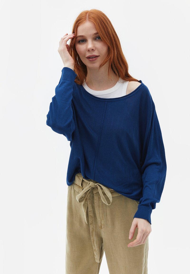 Blauwe oversized trui met brede halslijn en geribbelde manchetten, gecombineerd met beige hoge taillebroek die bij de taille is gestrikt.