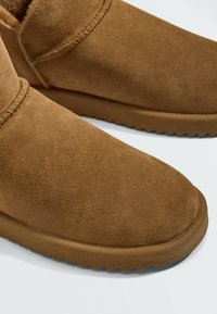 Gros plan sur des bottes d'hiver en daim marron avec des semelles épaisses en caoutchouc et des coutures visibles au niveau de la cheville sur fond blanc.