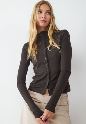 Jonge vrouw met lang blond haar die een donker, knoopsluitend, top met lange mouwen en een lichtbeige rok draagt, staand tegen een witte achtergrond.