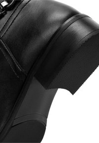 Botte en cuir noir avec une surface texturée, un talon incurvé, et une semelle en caoutchouc munie d'un motif antidérapant. Détails de couture en accent.