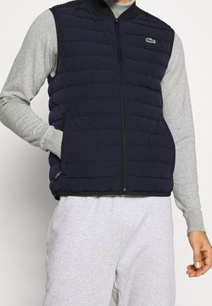 Man draagt een marineblauw doorgestikt mouwloos vest over een lichtgrijs overhemd met lange mouwen en lichtgrijze broek, hand in het vestzakje.