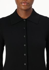 Top nero lavorato a maglia con chiusura a bottoni, caratterizzato da un colletto netto e una texture a coste. I bottoni sono neri con una finitura liscia.