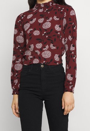 Vrouw draagt een langarmige bordeauxrode top met witte bloemenpatronen en zwarte broek met hoge taille, staand tegen een effen lichte achtergrond.