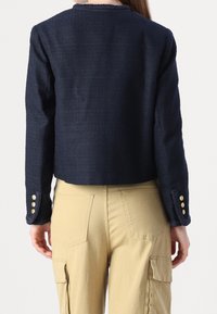 Navy blue textured jacket with a cropped design, featuring long sleeves and gold-button accents on the cuffs, worn over khaki pants.