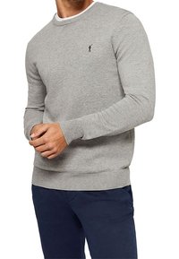 Homme portant un pull léger gris clair à col rond, ajusté, avec un petit logo noir brodé sur la poitrine, associé à un pantalon bleu foncé.
