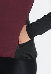 Gilet bordeaux avec coutures horizontales et une poche zippée, associé à des manches longues noires. Texture lisse et coupe ajustée.