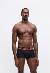 Boxer briefs neri con una ampia cintura rossa Calvin Klein, tessuto liscio, design aderente e lunghezza a metà coscia. Il modello si presenta con sicurezza.