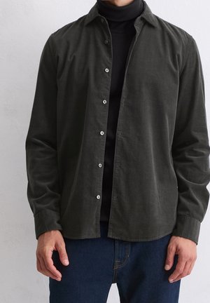 Mand iført mørke olivengrøn knap-op-skjorte over sort rullekrave og blå jeans, stående mod en ensartet lys baggrund.