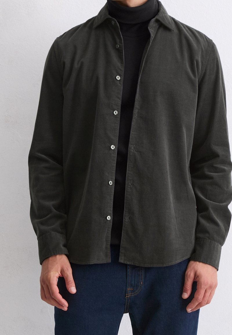 Homme portant une chemise boutonnée vert olive foncé sur un col roulé noir et un jean bleu, debout devant un fond clair uni.