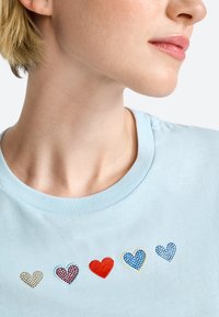 Šviesiai mėlynas medvilninis marškinėliai su penkiomis širdelėmis įvairių spalvų ir tekstūrų: dvi mėlynos, viena raudona, viena auksinė ir viena raštuota.