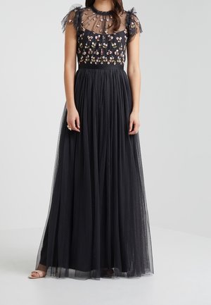 Kobieta w czarnej, długiej sukni do podłogi z przezroczystym górnym haftowanym kwiatowym motywem i krótkimi bufkami, stojąca na jednolitym tle.