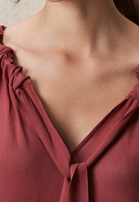 Blouse à col en V en tissu rose pâle avec détail de volant froncé aux épaules et col noué, texture lisse et apparence légère.