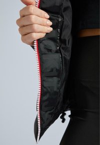 Ruka s světlou pletí a přírodními nehty drží otevřenou černou bundu s červeným a bílým zipem, ukazující vnitřní černou podšívku s velkým tmavým textem.