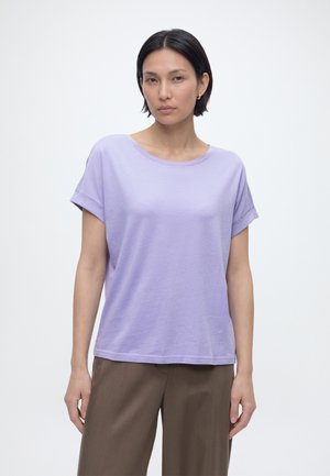 Γυναίκα με κοντά μαύρα μαλλιά, φοράει ανοιχτό μωβ κοντομάνικο πουκάμισο και καφέ παντελόνι, στέκεται μπροστά σε ένα απλό λευκό φόντο.