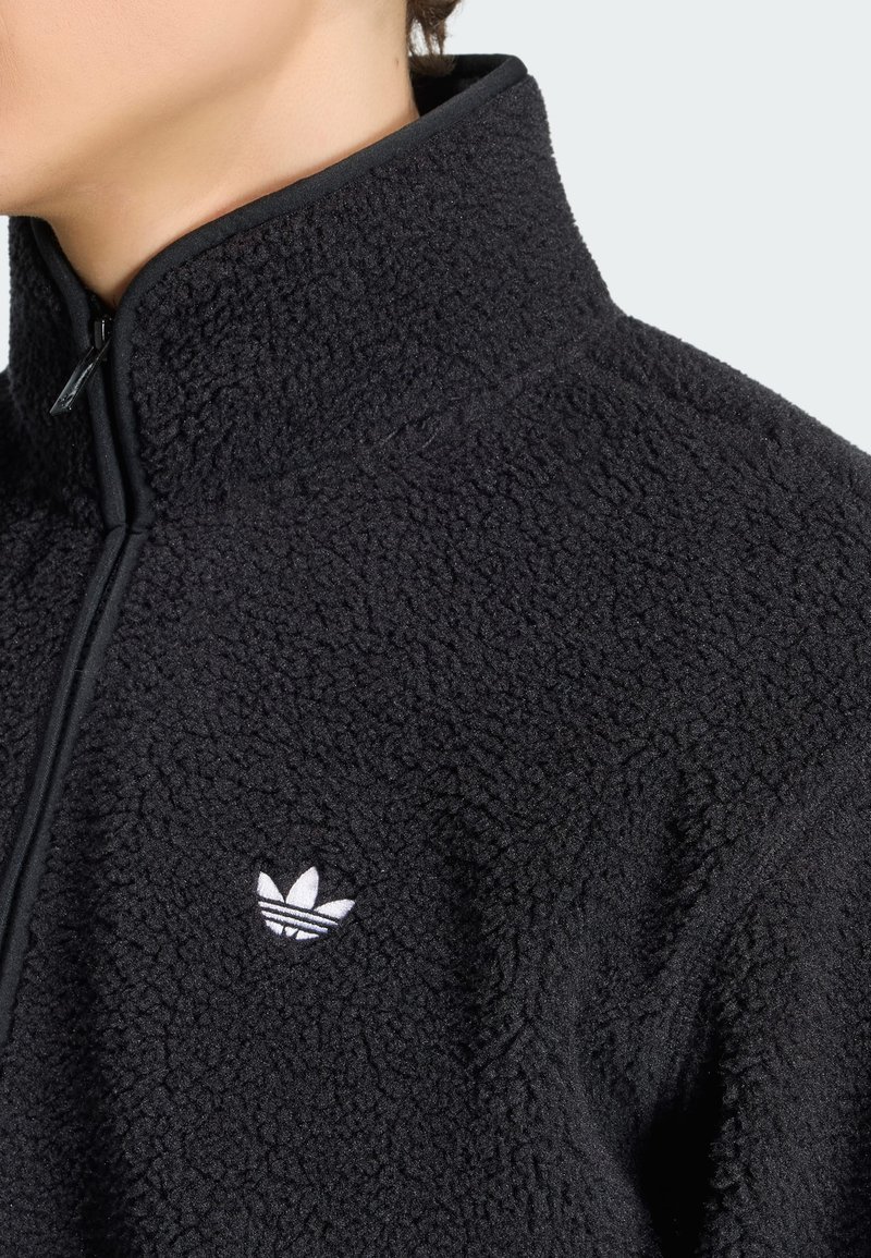Gros plan d'une personne portant une veste polaire noire texturée avec un col montant et un petit logo Adidas blanc sur la poitrine.