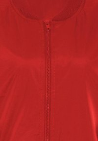 Röd zip-jacka med rund halsringning och slät textur. Framdelen har en dragkedjestängning som går från nacke till fåll, med en ribbad krage.
