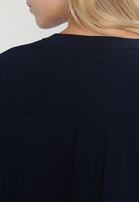 Vêtement bleu marine en tissu texturé, avec un col rond et un design plissé dans le dos, visible des épaules jusqu'à mi-dos.