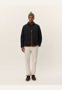 Giacca trapuntata di un blu scuro, con zip e bottoni a pressione, indossata sopra un maglione marrone, abbinata a pantaloni color crema e scarpe marroni. Berretto in testa.