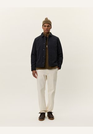 Chaqueta acolchada de un azul marino oscuro, con cremallera y botones a presión, llevada sobre un suéter marrón, combinada con pantalones de color crema y zapatos marrones. Gorro en la cabeza.