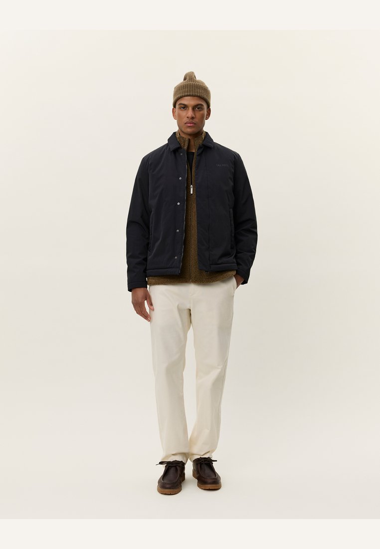 Chaqueta acolchada de un azul marino oscuro, con cremallera y botones a presión, llevada sobre un suéter marrón, combinada con pantalones de color crema y zapatos marrones. Gorro en la cabeza.