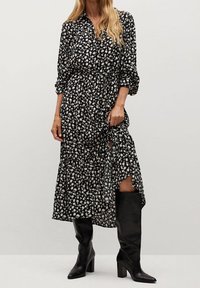 Schwarzes, blühendes Maxikleid mit durchgehendem Knopfverschluss, langen Ärmeln, tailliertem Bund und hohem Schlitz. Kombiniert mit knielangen schwarzen Stiefeln.
