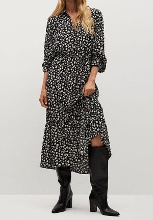Schwarzes, blühendes Maxikleid mit durchgehendem Knopfverschluss, langen Ärmeln, tailliertem Bund und hohem Schlitz. Kombiniert mit knielangen schwarzen Stiefeln.