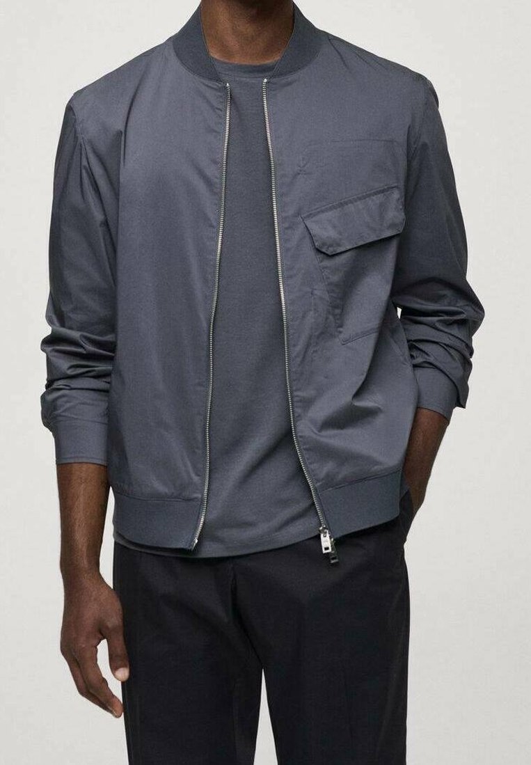 Homme portant une veste bomber bleu foncé sur un t-shirt assorti et un pantalon noir, debout avec une main dans la poche.