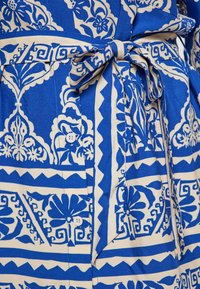 Tessuto patterned blu e beige con motivi floreali, un papillon in vita e un dettaglio con bottone. Il tessuto appare liscio con una leggera lucentezza.