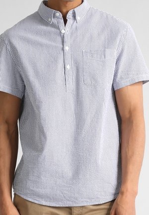 Homme portant une chemise blanche à manches courtes, boutonnée, avec de fines rayures verticales bleu marine et une poche poitrine, associée à un pantalon beige.