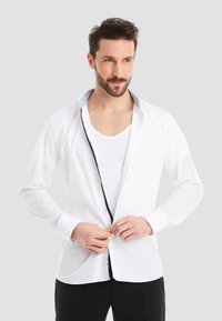 White button-up shirt with contrasting dark trim along the placket, long sleeves, and a collared neckline, worn over a white undershirt.
