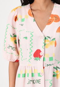 Ρόδινο φόρεμα με κοντά φουσκωτά μανίκια, V-λαιμόκοψη και σχέδια με φρούτα σε αντίθεση με πράσινες και κίτρινες λεπτομέρειες. Διακριτά ροζ κουμπιά.