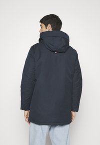 Navy blue insulated jacket with a large hood, zip closure, and elasticated cuffs, featuring a subtle brand logo on the back.