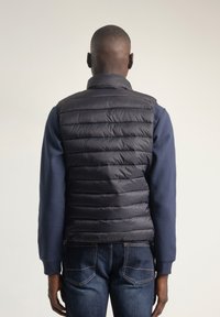 Chaleco acolchado negro con costuras horizontales, cuello alto y material ligero, llevado sobre una camiseta de manga larga azul marino.