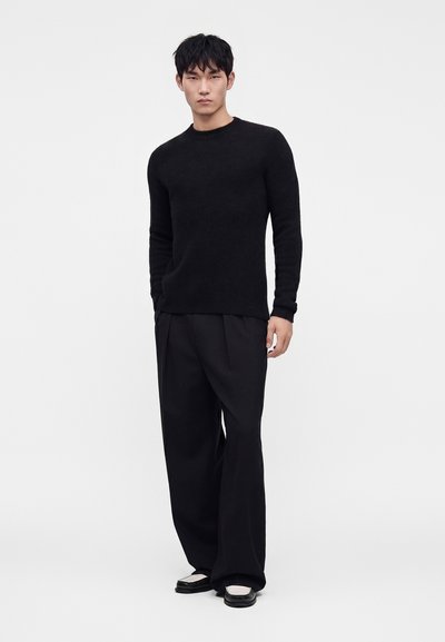Mann steht, trägt einen schwarzen langärmeligen Pullover, lockere schwarze Hose und schwarz-weiße Schuhe vor einem schlichten hellen Hintergrund.