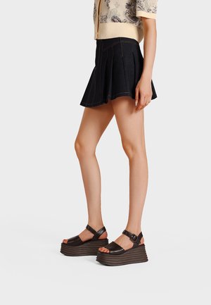 Donna con minigonna nera plissettata, top beige a maniche corte con fantasia e sandali con plateau neri con cinturini alla caviglia, in piedi di lato.