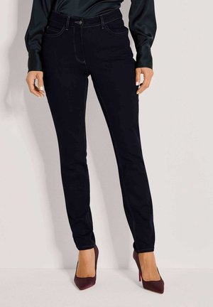 Frau trägt schwarze Slim-Fit-Jeans, dunkle Satinbluse und bordeauxrote spitze High Heels und steht vor einer weißen Wand.