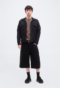 Νέος άνδρας στέκεται φορώντας μαύρο τζιν μπουφάν, καφέ μπλούζα με στρογγυλή λαιμόκοψη, μαύρες φαρδιές σορτς, μαύρες κάλτσες και μαύρα παπούτσια χωρίς κορδόνια.