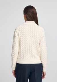 Sweter w kremowym kolorze z dzianiny w warkocze, z prążkowanymi mankietami i dolnym brzegiem. Pod spodem nosi jasną koszulę w kratę z kołnierzykiem, pokazany z tyłu.