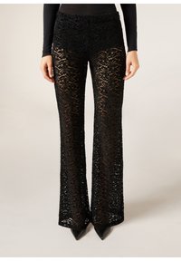Pantalon évasé en dentelle noire porté avec des chaussures noires à bout pointu et un haut noir à manches longues ajusté, dévoilant un motif floral en dentelle complexe et un design transparent.