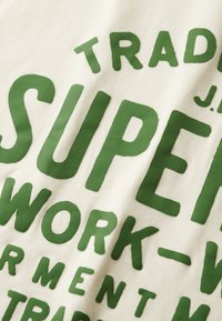 Tejido de algodón color crema con gráficos verdes llamativos. El texto incluye "SUPER" y "WORK" en grandes letras mayúsculas con un acabado texturizado.