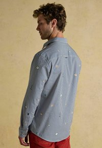 Pruhovaná košeľa s dlhým rukávom v modro-bielej farbe s farebnými vyšívanými vzormi. Obsahuje klasický golier a gombíkové manžety.