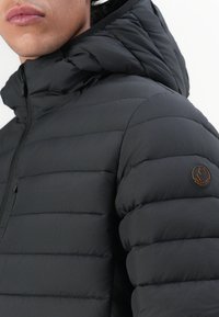 Sort puffjakke med vandret syede segmenter, en hætte og et rundt logo-patch på venstre skulder. Glat, struktureret stof.