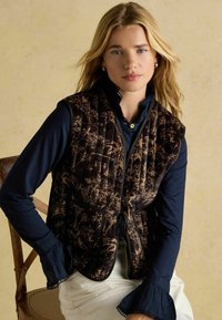 Prešívaná vesta tmavohnedej farby s kvetinovým vzorom; má vrecká, zapínanie na zaväzovanie a pod ňou je kontrastná jednofarebná tmavomodrá košeľa s dlhým rukávom.
