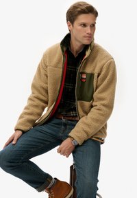 Beige fleecejas met groene accenten en rode ritsdetail, voorzien van een borstzak. Gedragen over een geruite blouse en gecombineerd met een blauwe spijkerbroek.