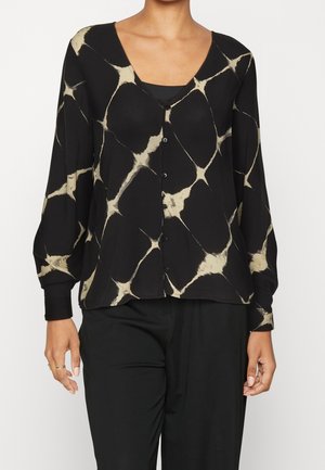 Kobieta ubrana w czarną bluzkę na guziki z beżowym abstrakcyjnym wzorem batiku i długimi rękawami, w połączeniu z czarnymi spodniami.