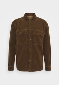 Chemise en velours marron à manches longues avec fermeture à boutons et deux poches avant à rabat, présentée sur un fond blanc uni.